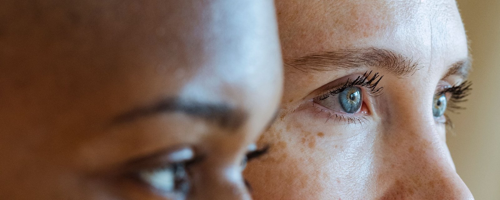 Two women in profile, one with deep brown skin and one with freckled fair skin, looking to the side