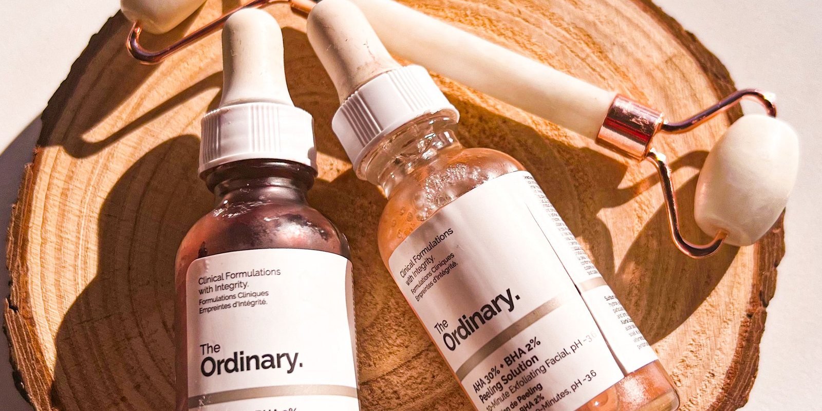 Two serum bottles and a rose quartz face roller arranged on a wooden slice