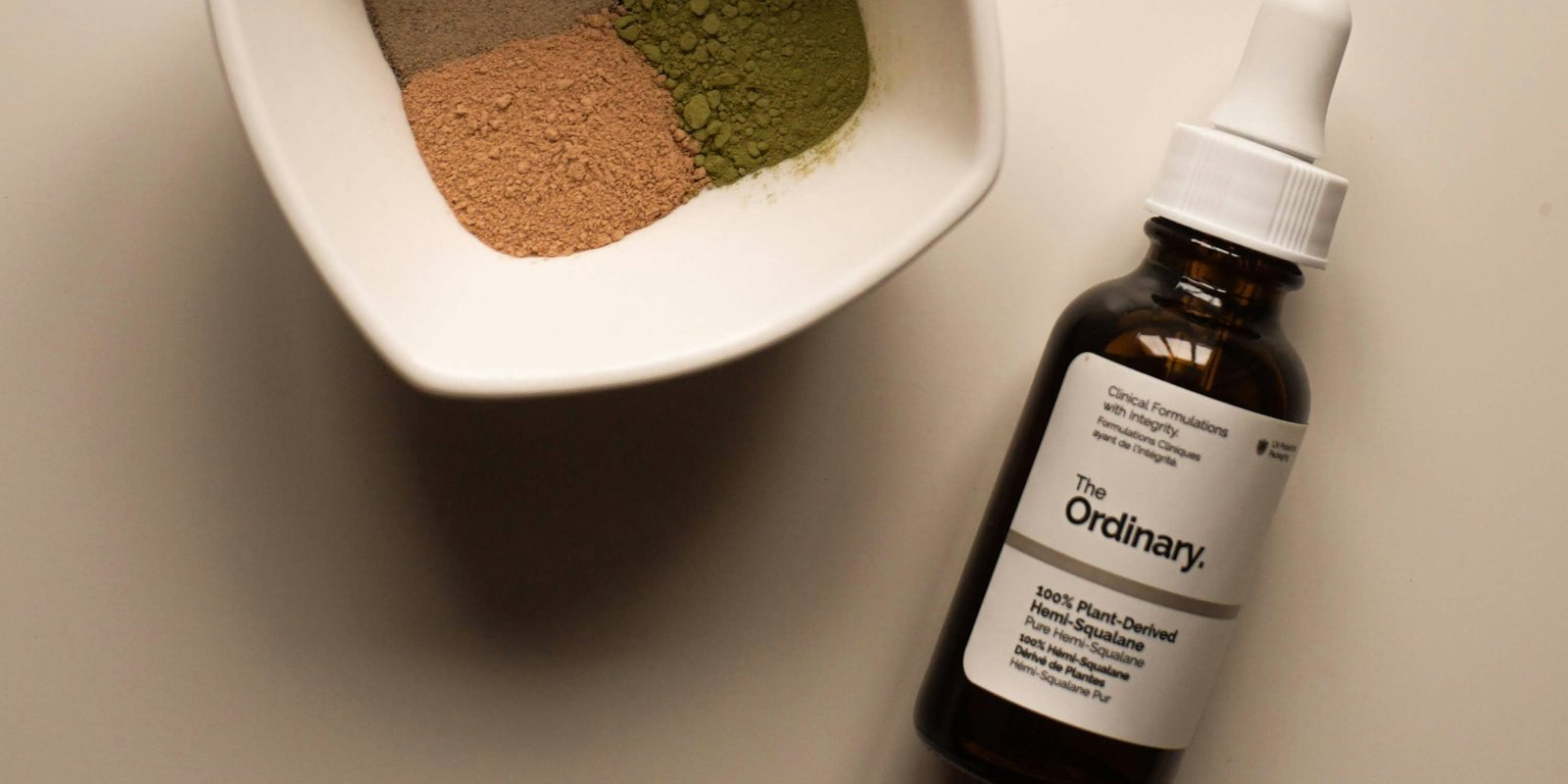 A single serum bottle beside a small ceramic dish of powders on a warm cream surface